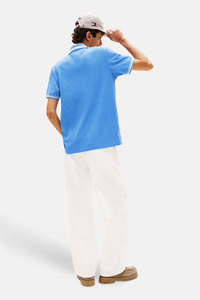 Polo bleu clair de Tommy Jeans, avec manches courtes à rayures blanches, porté avec un pantalon blanc et des chaussures marron.