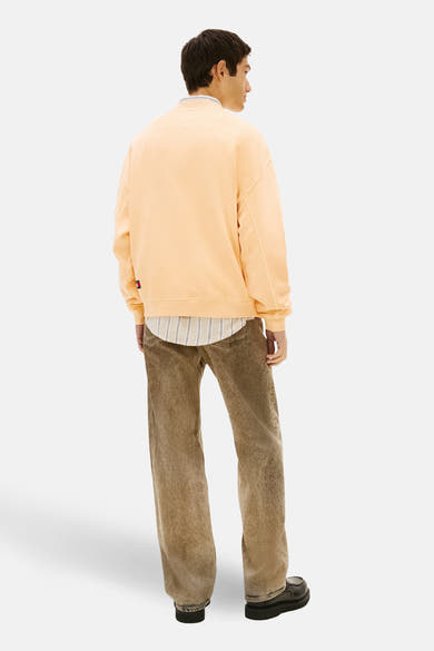Oranje sweatshirt van TOMMY JEANS, met een ronde hals en een relaxte pasvorm, gedragen over een gestreept overhemd en een bruine broek.