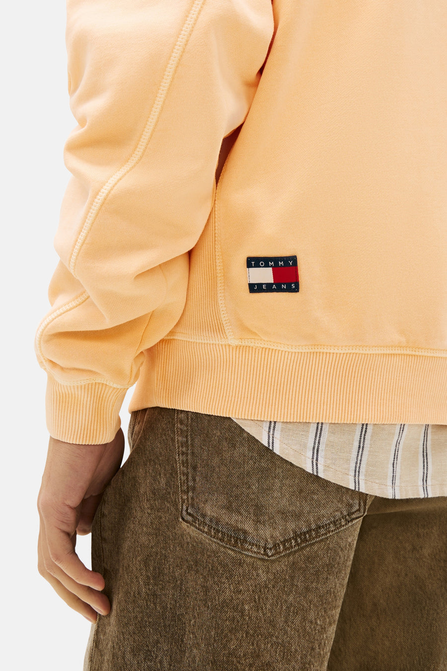 Oranje sweatshirt met ronde hals van TOMMY JEANS, geribbelde details, gedragen met een gestreept overhemd en een bruine broek.
