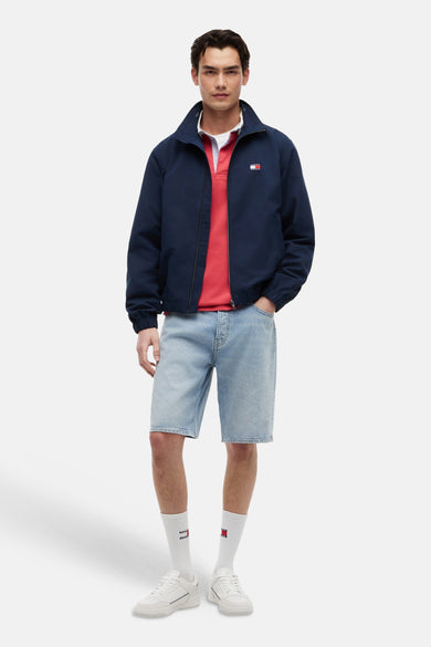 Blauwe regenjas van TOMMY JEANS, gedragen met een rode polo, een spijkerbroekshort en witte sneakers.