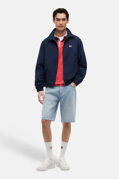 Blauwe regenjas van TOMMY JEANS, gedragen met een rode polo, een spijkerbroekshort en witte sneakers.