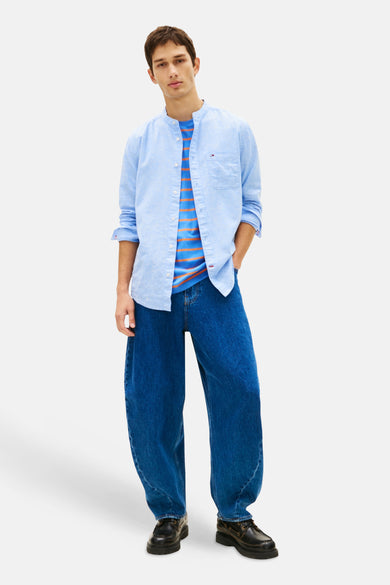Chemise bleu clair à manches longues de TOMMY JEANS, portée ouverte sur un t-shirt rayé, avec un jean ample.
