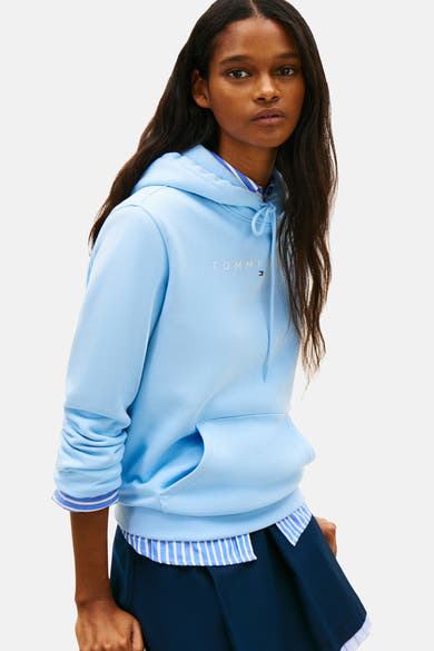 Blauwe hoodie van Tommy Jeans, gedragen over een gestreept overhemd, met discreet logo op de voorkant.