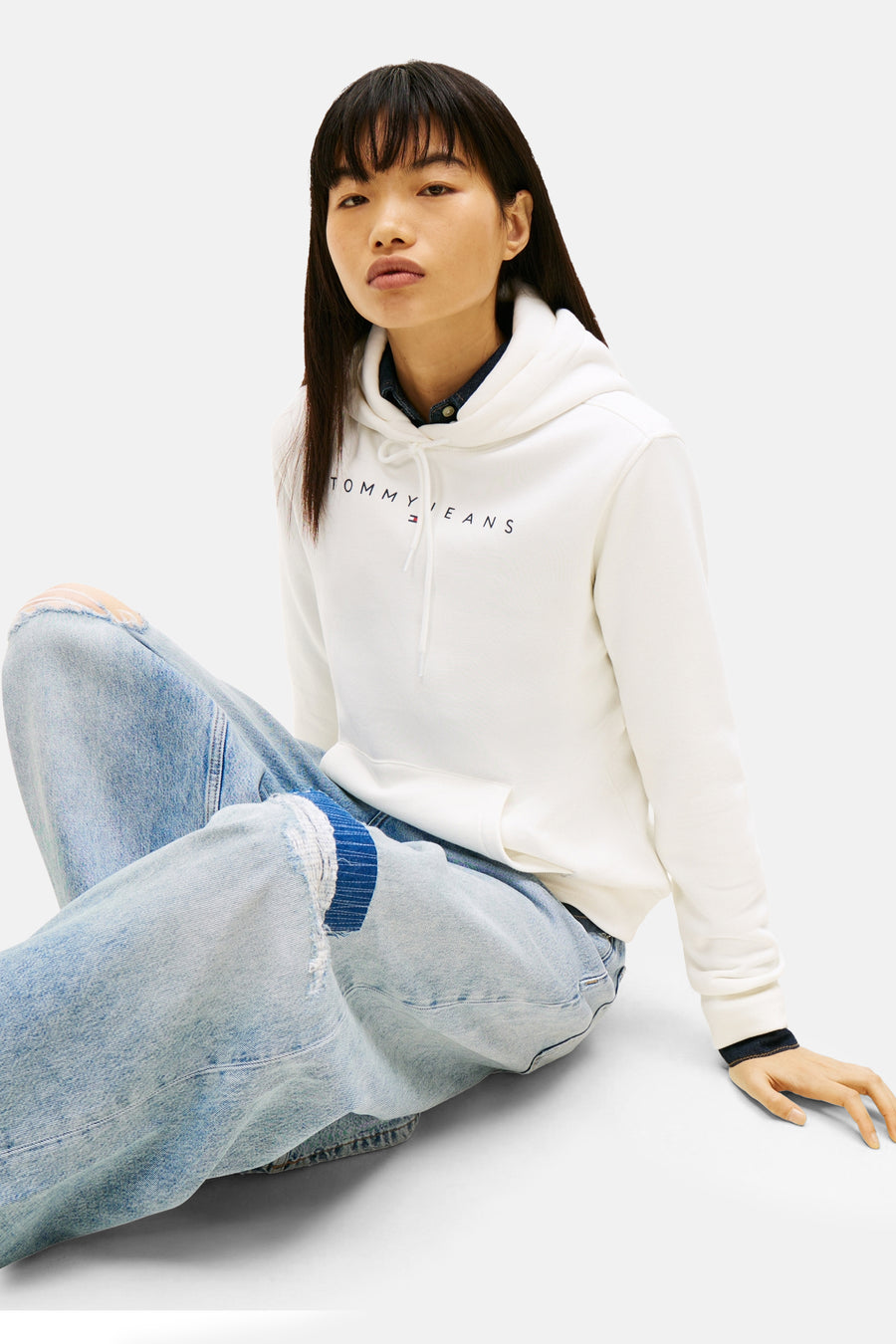 Witte hoodie van TOMMY JEANS, gedragen met een lichtblauwe gescheurde jeans en een donkerblauwe blouse.