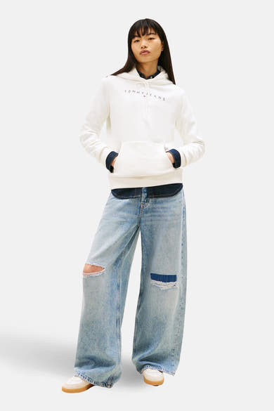 Witte hoodie van TOMMY JEANS, met een opdruk van het logo, gedragen met een marineblauw overhemd en een wijde, gescheurde spijkerbroek.