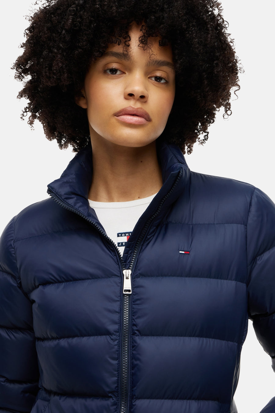 Navy blue quilted puffer jacket by Tommy Hilfiger, front view with stand collar and silver zipper, worn over white ribbed top.