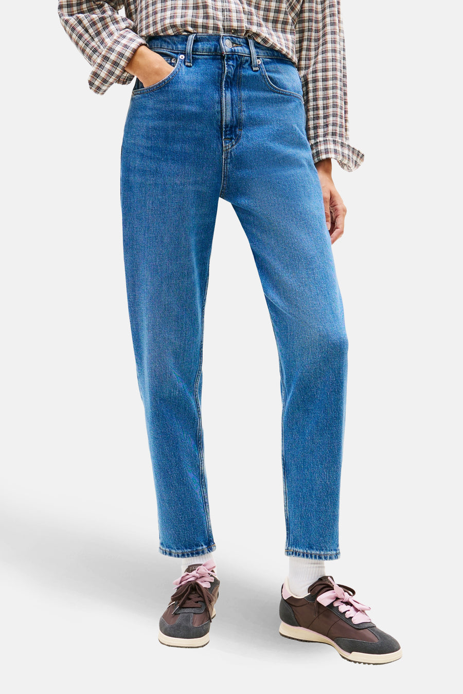 Jean Mom en denim bleu moyen de TOMMY JEANS, coupe décontractée, porté avec une chemise à carreaux et des baskets marron.
