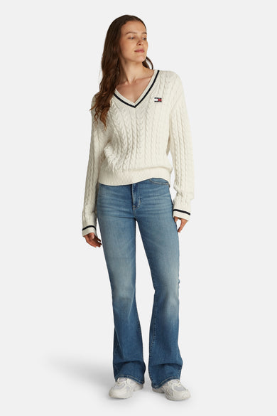 Pull à col en V blanc de Tommy Jeans, avec motif torsadé et rayures contrastantes au col et aux poignets, porté avec un jean bleu.
