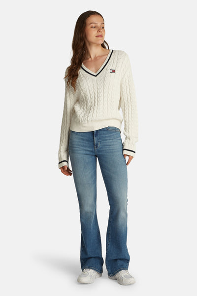 Pull à col en V blanc de Tommy Jeans, avec motif torsadé et rayures contrastantes au col et aux poignets, porté avec un jean bleu.
