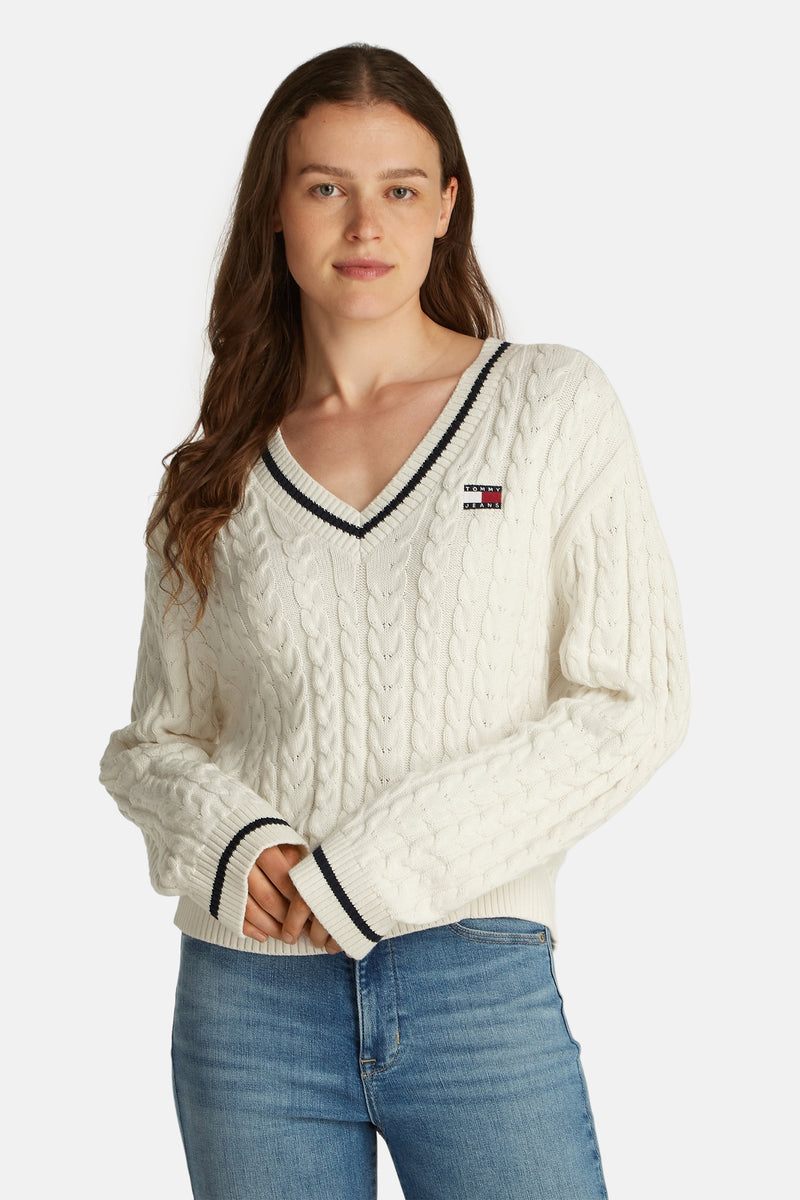 Pull à col en V blanc de TOMMY JEANS en maille torsadée avec finitions rayées et porté avec un jean.
