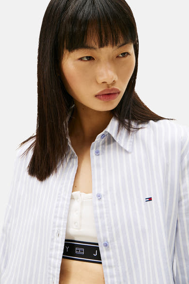 Blauwe gestreepte blouse met lange mouwen van TOMMY JEANS, gedragen open over een witte tanktop en een logoband.