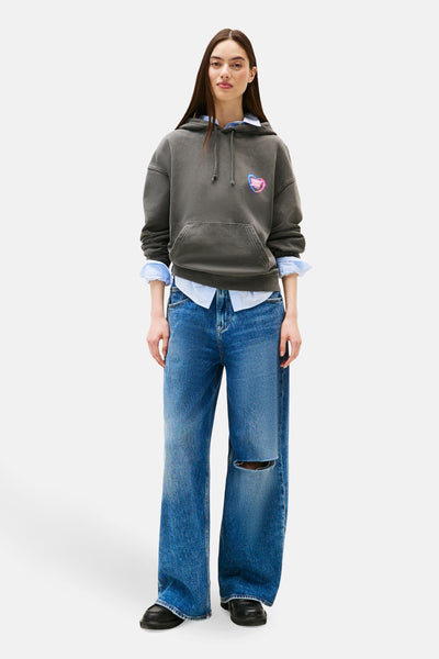 Grijze Tommy Jeans hoodie met neon hartpatroon, gedragen over een blauw overhemd en een gescheurde spijkerbroek.