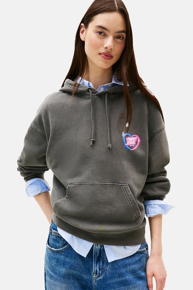 Grijze hoodie van TOMMY JEANS met neon hartlogo, gedragen over een blauw overhemd en een spijkerbroek.