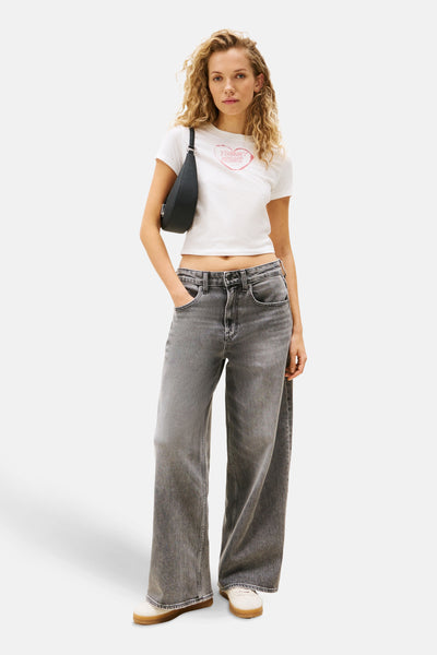 Wit T-shirt met korte mouwen van TOMMY JEANS, gedragen met een grijze high-waist jeans en een zwarte schoudertas.
