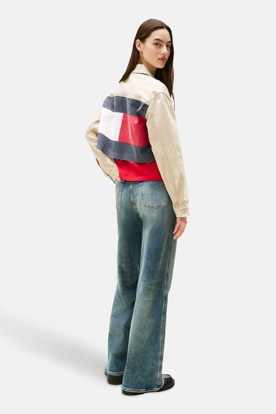 Korte beige jas van TOMMY JEANS, versierd met een grote kleurrijke vlag op de rug, gedragen met een rood T-shirt en een wijde jeans.