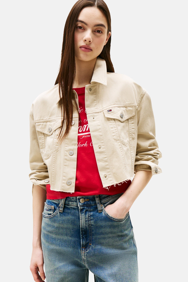 Korte beige jas van TOMMY JEANS met metalen knopen, gedragen over een rood T-shirt en een blauwe jeans.