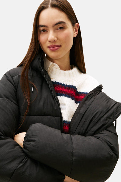 Veste courte noire par TOMMY JEANS avec une texture matelassée, portée sur un pull rayé blanc et rouge.