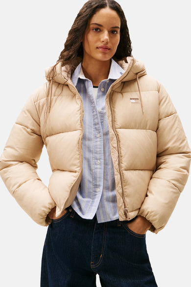 Korte beige jas van TOMMY JEANS, met capuchon en ritssluiting, gedragen over een blauw gestreept overhemd en een donkere spijkerbroek.