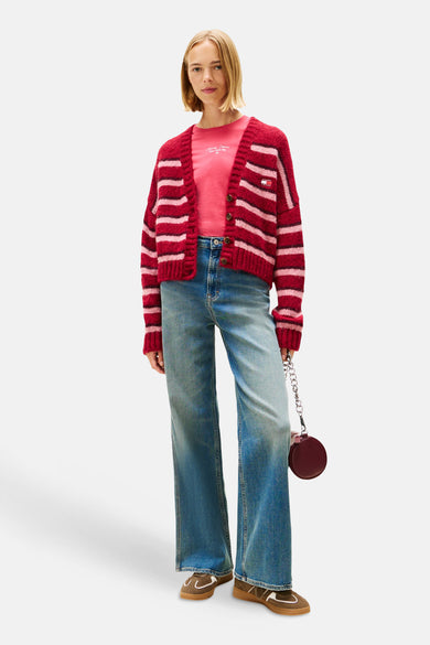 Gilet rouge à rayures par TOMMY JEANS, porté sur un t-shirt rose, avec un jean bleu ample et des chaussures marron.
