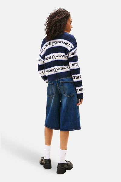TOMMY JEANS blauwe V-hals trui met strepen en logo, gedragen met een spijkerbroek short en zwarte schoenen.