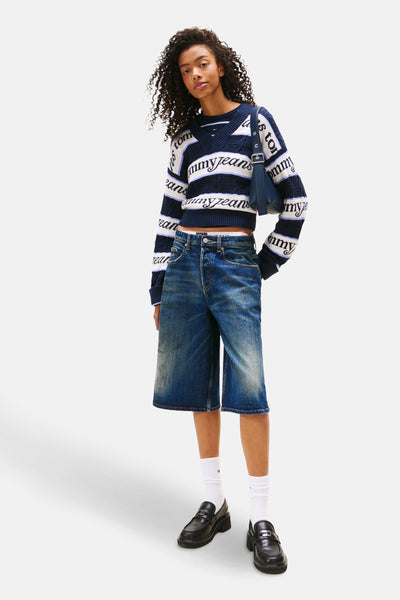 Blauwe V-hals trui van TOMMY JEANS, gestreept met logo op de mouwen, gedragen met een spijkershort en loafers.