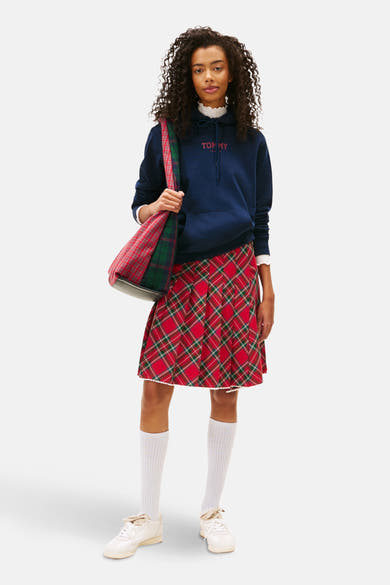 Sweat à capuche bleu de TOMMY JEANS avec logo rouge, porté avec une jupe à carreaux, des chaussettes blanches et des baskets.

