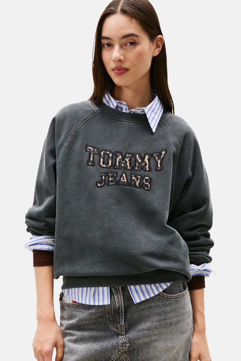 Grijze rondehals sweatshirt van TOMMY JEANS, met luipaardprint "TOMMY JEANS" motief, gedragen over een gestreept overhemd en een grijze jeans.