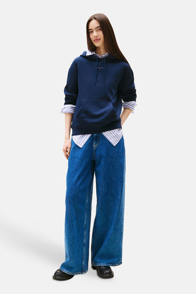 Blauwe hoodie van TOMMY JEANS, gedragen over een gestreept overhemd en met een wijde spijkerbroek.