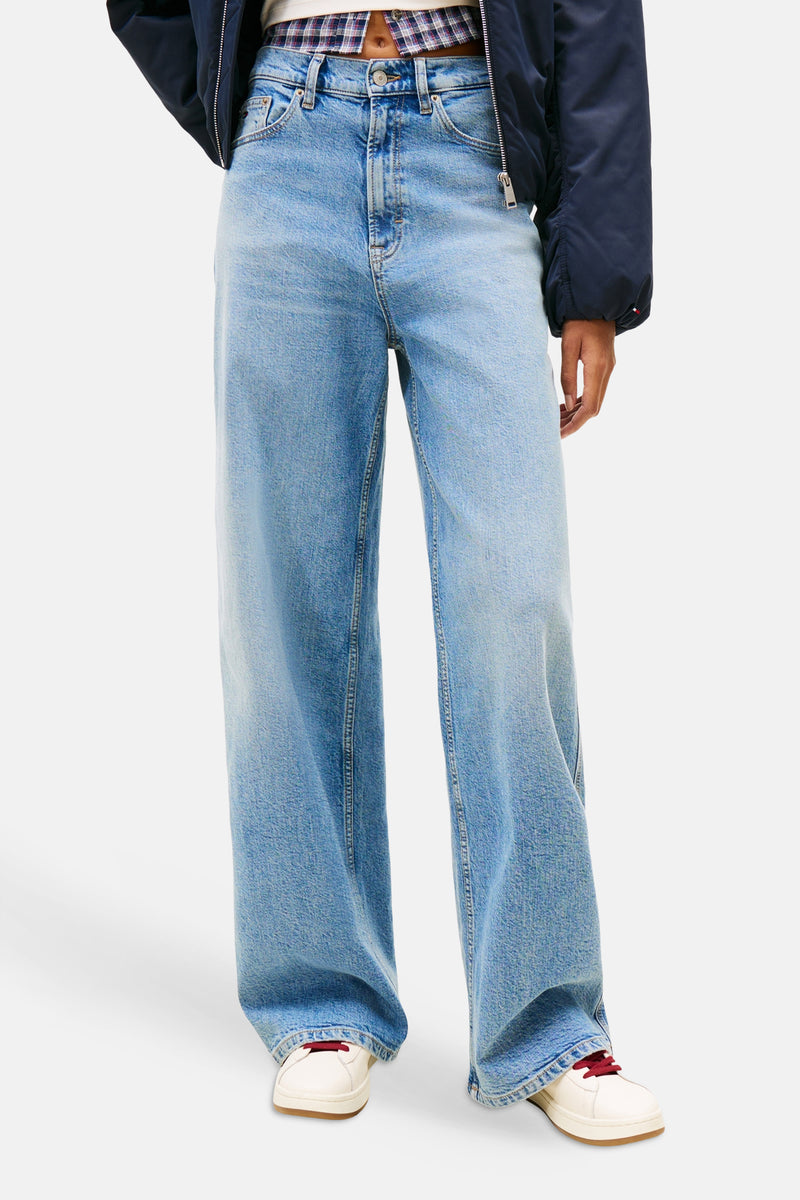 Jean large en denim bleu moyen de TOMMY JEANS, porté avec une veste bleu marine et des chaussures de sport crème.
