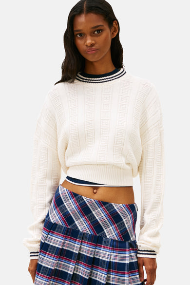 Pull à col rond blanc de TOMMY JEANS, avec motif texturé et bordures rayées, porté avec une jupe à carreaux.
