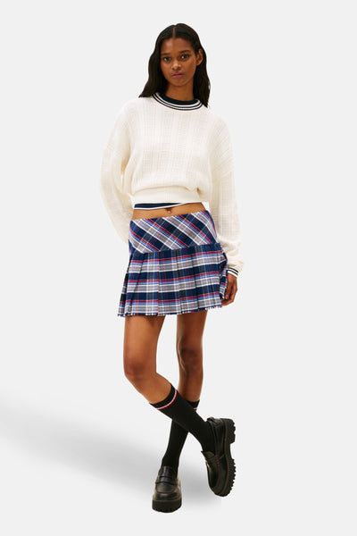 Pull à col rond blanc de TOMMY JEANS, avec col et poignets rayés, porté avec une jupe plissée à carreaux.
