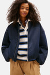 Veste courte bleue de TOMMY JEANS avec fermeture éclair argentée, portée sur un polo rayé et un chemisier à carreaux.