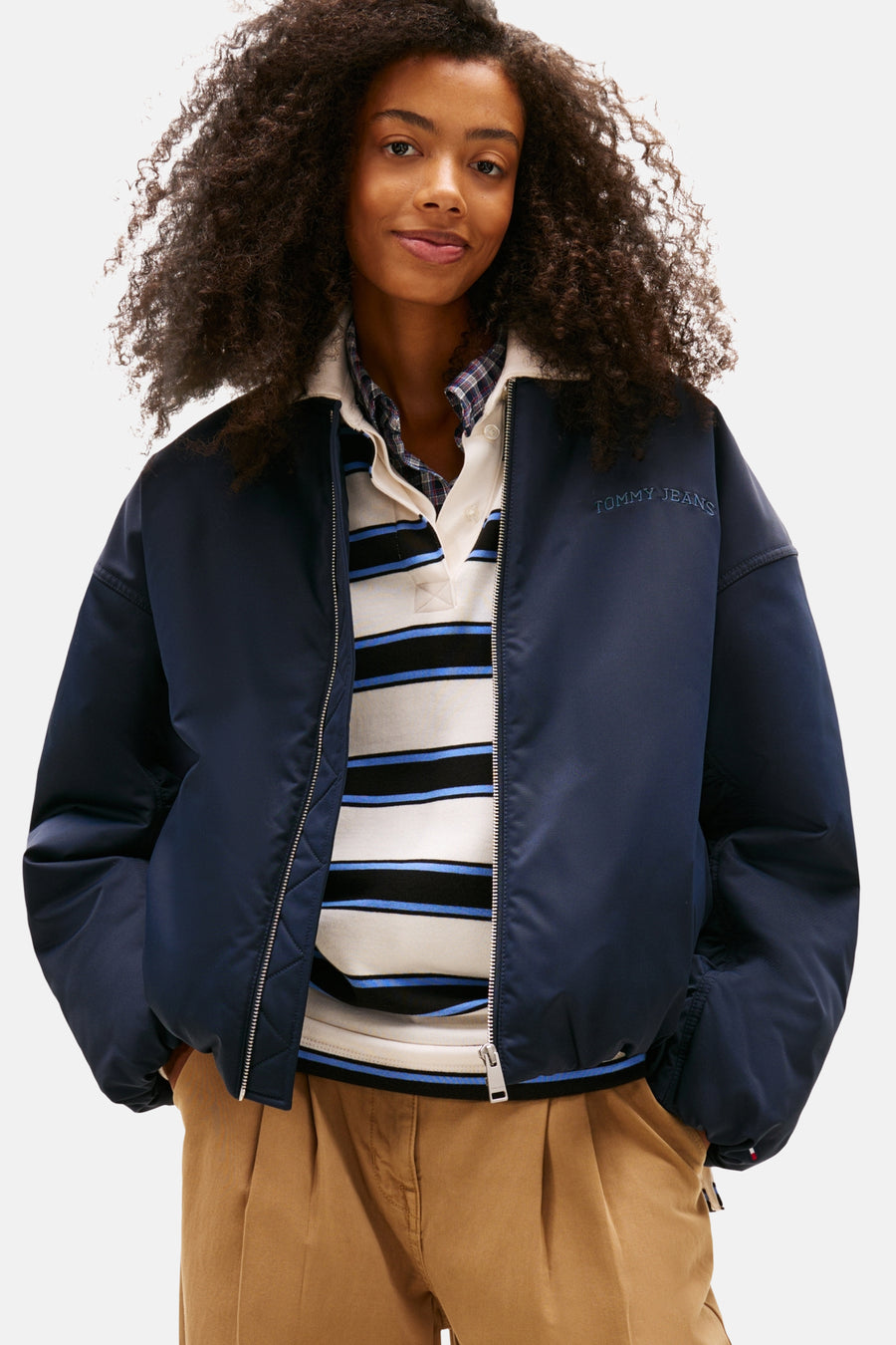 Veste courte bleue de TOMMY JEANS avec fermeture éclair argentée, portée sur un polo rayé et un chemisier à carreaux.