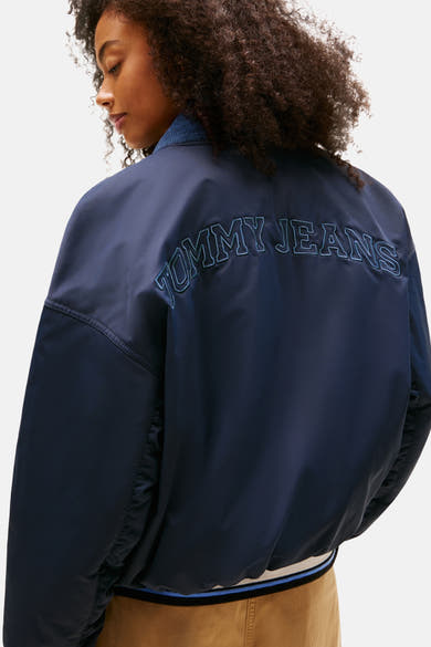 Veste courte bleue par TOMMY JEANS, avec logo brodé au dos, portée avec un pantalon beige.