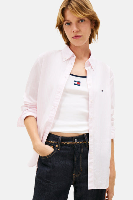 Chemise à manches longues rose de Tommy Jeans, portée ouverte sur un débardeur blanc et un jean taille basse avec ceinture chaîne.
