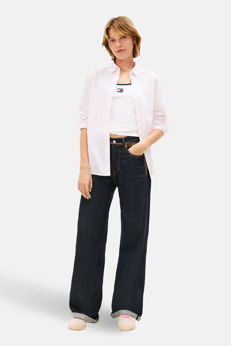Chemise rose à manches longues de Tommy Jeans, portée ouverte sur un débardeur blanc et un jean taille haute bleu foncé.
