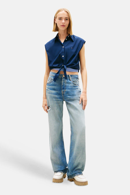 Chemisier bleu sans manches de TOMMY JEANS, noué à la taille, porté avec un jean ample et des chaussures à plateforme.
