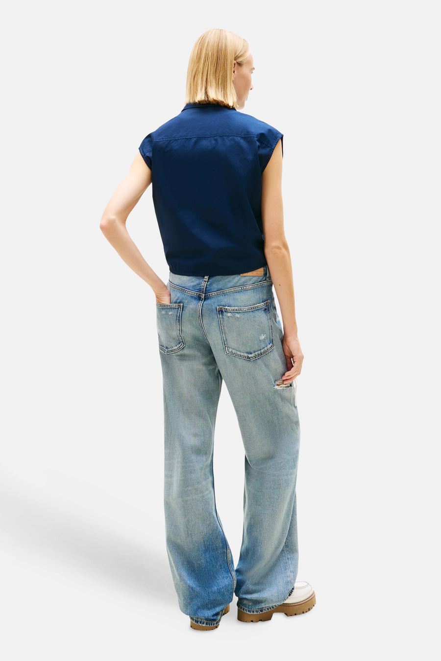 Blauwe blouse van TOMMY JEANS, mouwloos model, gedragen met een wijde, uitgewassen spijkerbroek en schoenen met dikke zool.