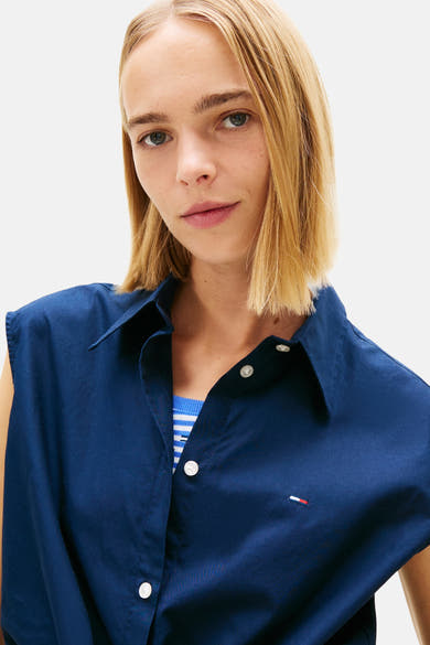 Blauw mouwloos overhemd van Tommy Jeans, gedragen over een gestreept hemdje, met discreet logo op de borst.