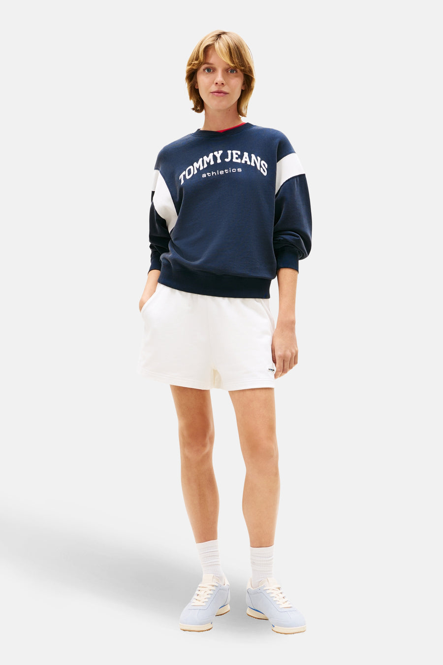 Short blanc de Tommy Jeans, porté avec un pull bleu marine à logo, des chaussettes blanches et des baskets bleues.
