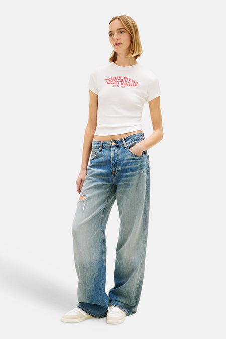 T-shirt blanc à manches courtes de TOMMY JEANS, avec un logo rouge sur le devant, porté avec un jean ample bleu.

