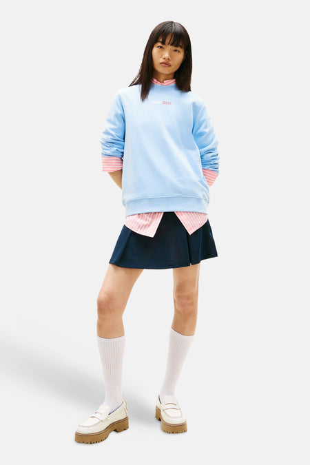 Sweat bleu de TOMMY JEANS, porté sur une chemise rayée rose et une jupe plissée bleu marine.
