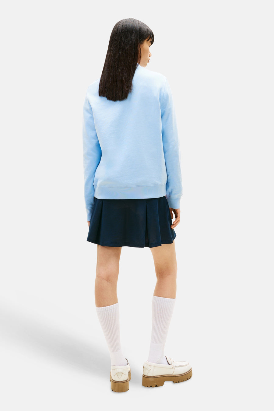Sweat-shirt bleu clair de Tommy Jeans, avec manches longues, porté avec une jupe bleu marine et des chaussures plateforme blanches.
