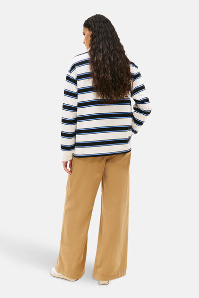 Polo à manches longues multicolore de TOMMY JEANS, rayures horizontales noires et bleues sur fond blanc, porté avec un pantalon beige.
