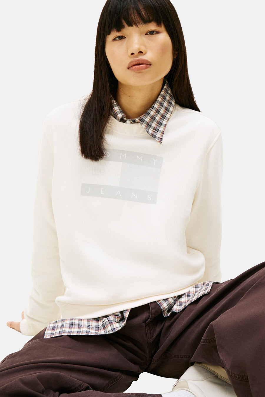 Ecru sweatshirt van TOMMY JEANS, gedragen over een geruit overhemd, met een bruine broek.