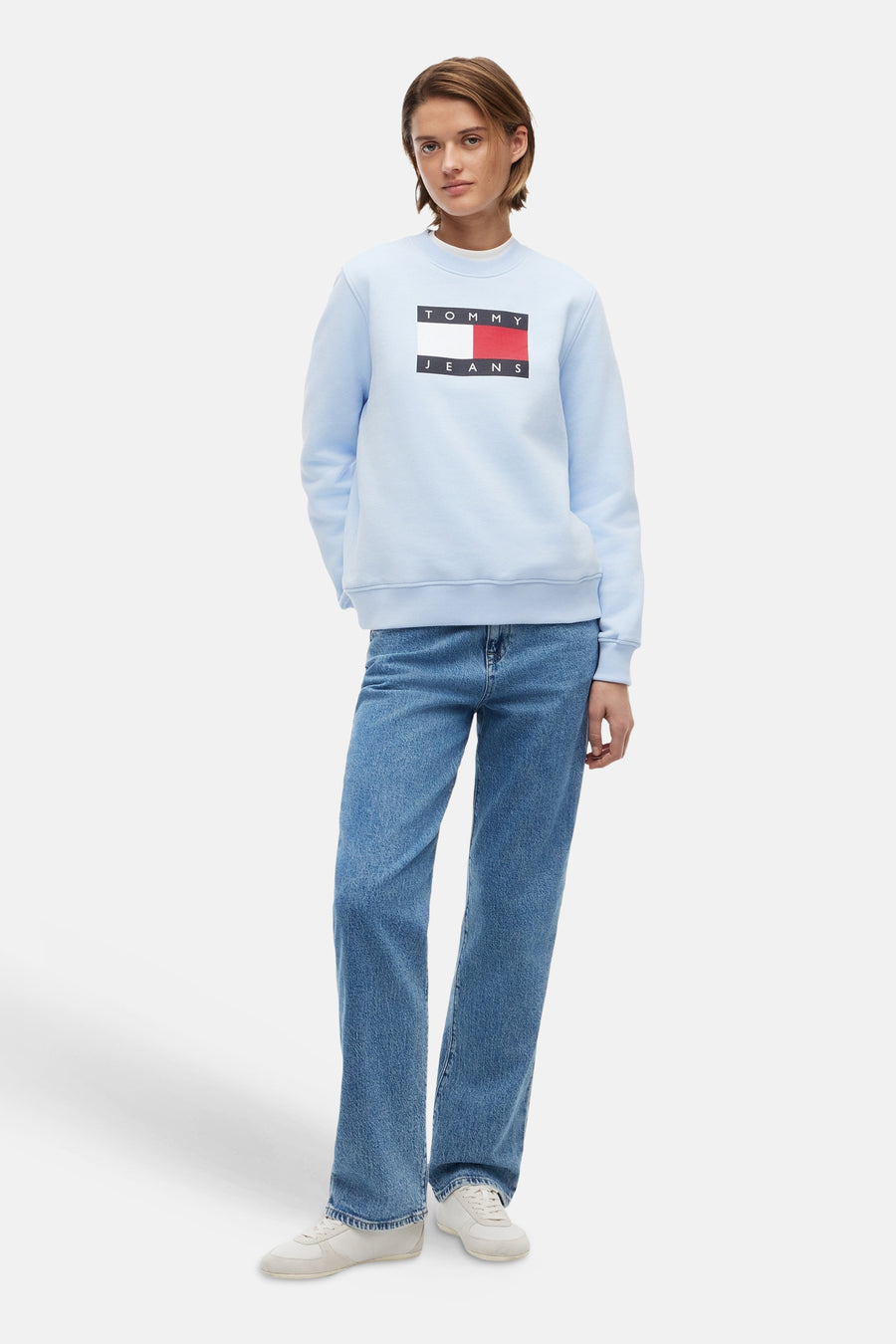 Lichtblauwe rondehals sweatshirt van TOMMY JEANS met een groot rechthoekig logo, gedragen met een spijkerbroek en witte sneakers.