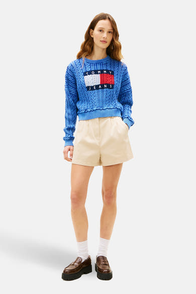 Short beige de TOMMY JEANS, porté avec un pull bleu à motif torsadé, des chaussettes blanches et des mocassins marron.