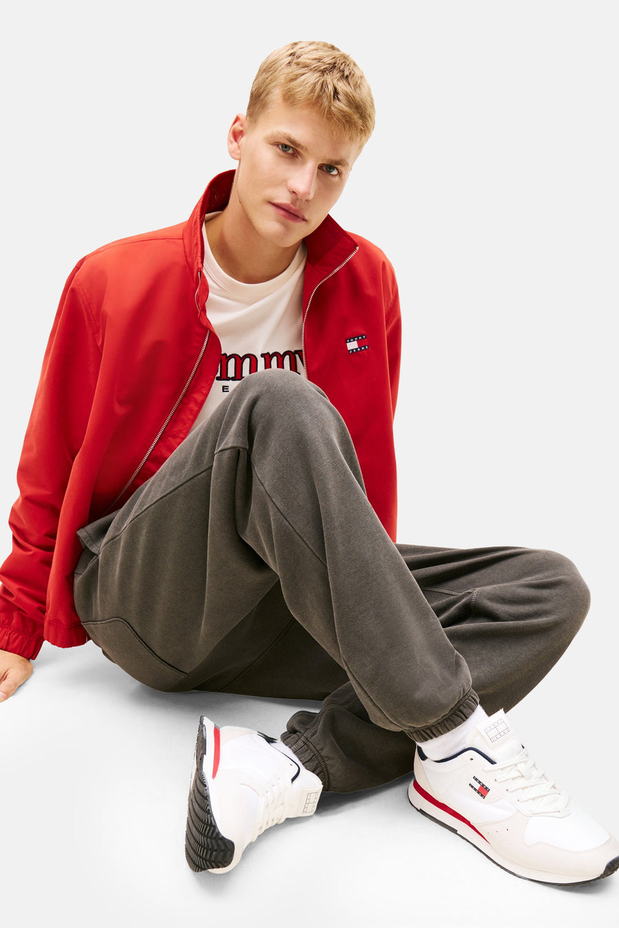 Witte Tommy Jeans sneakers, met een rode streep en gedragen met een grijze joggingbroek en een rood jack.