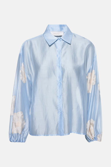 Chemise à manches longues bleu clair de AMELIE ET AMELIE, avec des motifs floraux brodés sur les manches.
