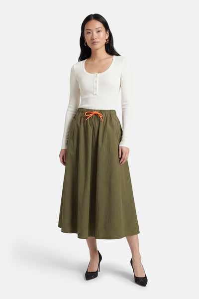 Jupe kaki 3/4 de Le Temps des Cerises, avec cordon de serrage orange, portée avec un haut blanc à manches longues et des escarpins noirs.
